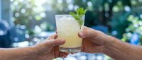 Hands holding refreshing lemonade or mojito cocktail with mint garnish outdoors in summer sunlight, perfect for beverage, hospitality, and lifestyle marketing.