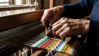 Skilled artisan hands weaving colorful threads on traditional wooden loom, creating handmade textile with vibrant rainbow pattern in workshop setting.