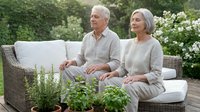Senior Caucasian couple relaxing together on outdoor patio furniture surrounded by lush garden plants and white flowers in peaceful backyard setting.