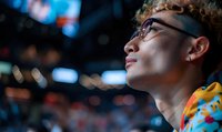 Young Asian male esports fan in glasses watching competitive gaming event with crowd in background, illuminated by arena lights and digital displays.