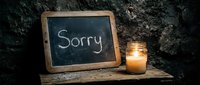 Chalkboard with the word Sorry beside a glowing candle in a glass jar on a rustic wooden shelf against a dark stone wall.