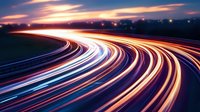 Long exposure shot of curving highway at dusk with colorful light trails from moving vehicles creating dynamic streaks against dramatic sunset sky.