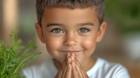 Young boy with bright blue eyes and tanned skin wearing white t-shirt praying or showing gratitude, green plant in foreground, blurred background creates peaceful mood.