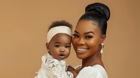 Happy African American mother holding adorable baby daughter against warm beige background, both wearing white outfits for family portrait session.