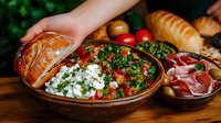 Mediterranean appetizer platter with fresh feta cheese, tomato salad, prosciutto, olives, and crusty bread served on rustic wooden table for party gathering.