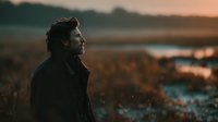 Young man with curly hair gazing at sunset over coastal landscape, contemplative mood, golden hour lighting for lifestyle and travel content.