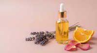 Golden essential oil serum bottle with dropper, dried lavender sprigs, orange slice and rose petals on pink background for skincare and aromatherapy.