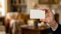 Senior man with light skin tone holding blank white business card in hand against blurred home interior background for branding and corporate identity mockup purposes.
