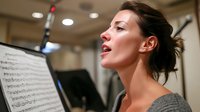 Female vocalist performing in recording studio, reading sheet music during professional session with focused expression and casual attire.