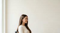 Young woman with long dark hair wearing casual beige top, standing indoors against neutral wall with natural light, suitable for lifestyle and wellness marketing materials.