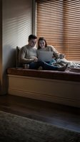 Happy young couple sitting together on window seat using laptop computer in cozy home interior with warm sunlight streaming through wooden blinds.