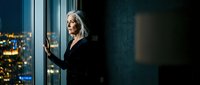 Elegant senior Caucasian businesswoman in black dress gazing at city lights from office window at night, reflecting on success.