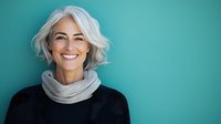 Mature woman with silver gray hair and bright genuine smile wearing black sweater and gray scarf against turquoise wall background, expressing joy and confidence.