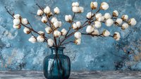 Cotton branches with fluffy white bolls arranged in blue glass vase against textured turquoise wall background, rustic farmhouse style home decor composition.