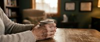 Elderly hands holding warm coffee mug on wooden table in cozy home interior, morning comfort and relaxation concept for seniors lifestyle.