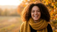 Young African American woman with curly hair laughing joyfully in autumn park wearing cozy yellow scarf during golden hour sunset for lifestyle content.