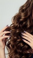 Woman with luxurious brown curly hair touching locks with manicured purple nails against neutral background for beauty and haircare concepts.