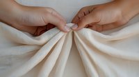 Close-up of hands pinching and gathering cream-colored fabric, creating elegant pleats and folds against a neutral background.