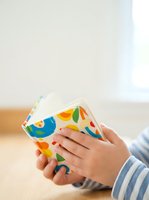 Toddler with light skin tone holding colorful fabric book with fruit patterns, wearing blue striped clothing. Educational toy for early childhood development and learning.