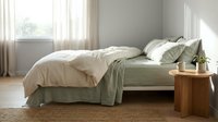 Serene bedroom interior with unmade bed featuring sage green and cream linens, wooden nightstand with plant, natural light from window, minimalist Scandinavian design aesthetic.