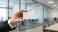 Businessman in formal suit holding blank white business card in modern office environment with glass partitions and blurred colleagues in background.