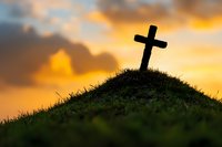 Christian cross silhouette on grassy hill against dramatic orange sunset sky with clouds, spiritual and religious concept for faith-based publications.