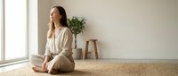 Young woman practicing mindfulness meditation sitting cross-legged on jute mat in minimalist room with natural light for wellness and mental health.