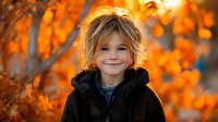 Adorable young child with tousled blonde hair wearing cozy black hoodie smiling warmly outdoors during golden autumn season with vibrant orange foliage backdrop.