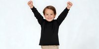 Cheerful young Caucasian boy with arms raised in victory pose wearing black long sleeve shirt against white background, expressing joy and achievement.
