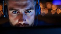 Young Caucasian man with beard wearing headphones in dark room illuminated by blue light and bokeh effects, focused intense gaze at computer screen, dramatic portrait.