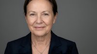 Mature woman with light skin tone and dark hair wearing dark blazer against neutral gray background, professional headshot.