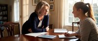 Professional middle-aged Caucasian female advisor consulting with young adult woman at wooden table in bright home office setting for business meeting.