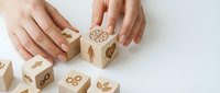 Hands arranging wooden blocks with leaf and gear icons on white surface, representing business growth, sustainability, and strategic planning for corporate design projects.