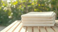 Fresh stack of folded white cotton towels on wooden surface outdoors with soft natural bokeh background, suitable for spa, wellness, and lifestyle designs.