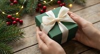 Hands holding green gift box with white ribbon bow surrounded by festive pine branches and red berries on rustic wooden background for holiday celebrations.