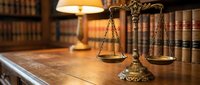 Scales of justice on wooden desk in law library with legal books and warm lamp lighting creating professional attorney office atmosphere.