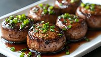 Grilled mushroom caps glazed with savory sauce and garnished with fresh chopped green onions on white rectangular serving plate, close up view of gourmet appetizer.