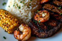 Grilled surf and turf dinner with juicy steak, seasoned shrimp, steamed white rice, and charred corn on the cob garnished with fresh herbs and butter.