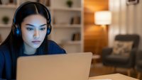 Young Asian woman wearing headphones working on laptop computer at home office desk in evening. Remote work, online learning, gaming, video call concept.