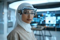 Female scientist in hijab wearing safety glasses and hard hat in modern laboratory with digital displays in background.