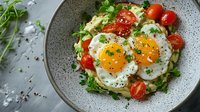 Fried eggs with fresh greens, tomatoes and herbs on speckled plate, ready for breakfast, brunch or healthy meal preparation.