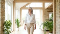 Mature woman with gray hair wearing white button-up shirt and beige pants walks through bright modern hallway with potted plants and wooden beams, suitable for lifestyle and business content.