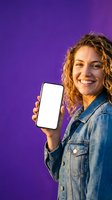Happy young woman with curly hair holding smartphone with blank screen on purple background for mobile app mockup and digital marketing campaigns.