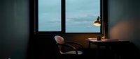 Minimalist workspace with wooden desk, desk lamp, and comfortable chair positioned by large windows overlooking cloudy sky, ideal for office and productivity concepts.