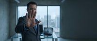 Man in dark business suit making stop gesture with hand raised toward camera in modern office with city skyline view through windows, suitable for corporate, rejection, and professional communication concepts.