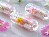 Clear capsules filled with pink powder and golden glitter surrounded by delicate flower petals on white surface, creating dreamy pharmaceutical aesthetic.