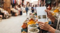 Vendor holding white pouch of spices at outdoor market stall with blurred crowd and merchandise in background, suitable for commerce, retail, and food trade concepts.
