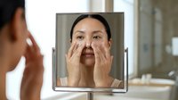 Woman with East Asian ethnicity examining facial skin in mirror, touching cheeks and nose area, appearing concerned about complexion or blemishes in bathroom setting.