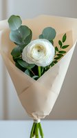 Single white ranunculus flower wrapped in minimal cream paper with eucalyptus leaves and greenery, creating an elegant modern bouquet for weddings and special occasions.
