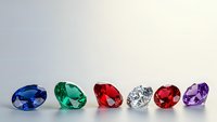 Colorful collection of precious gemstones including sapphire, emerald, ruby, diamond and amethyst arranged in elegant row on neutral background.